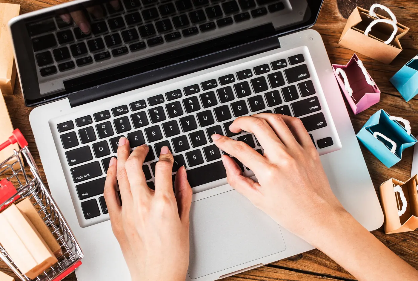 Overhead view of hands typing on a laptop surrounded by miniature shopping bags and a small trolley
