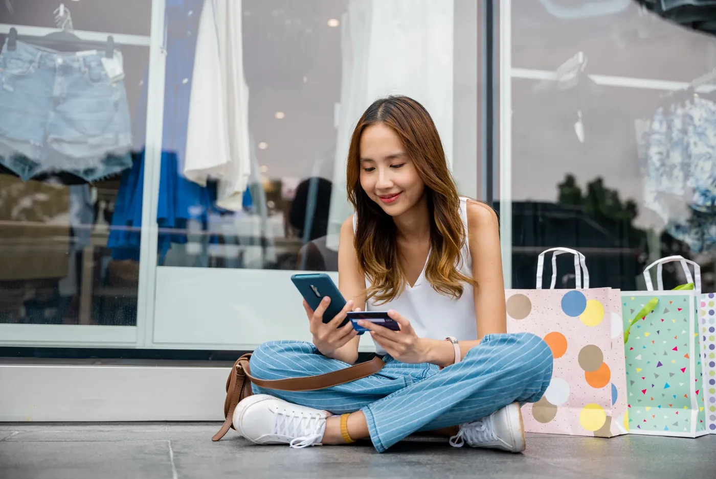 A person sitting outside a shop looking at a phone while holding a payment card